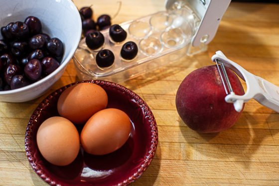 small bowl of cherries, cherry pitter, 3 brown eggs, peach with peeler on cutting board