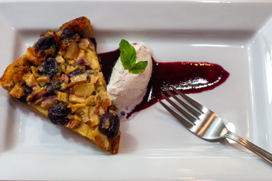white plate with wedge of clafoutis, drizzle of cherry sauce, vanilla ice cream and mint,fork on side