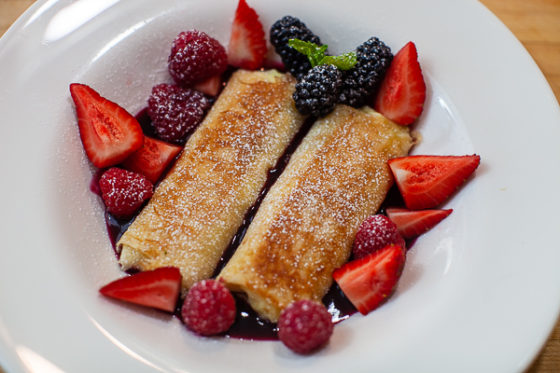 Bowl of two blintzes in cherry syrup surrounded by berries