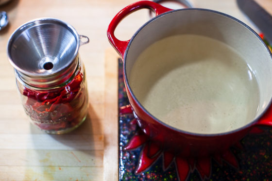 metal funnel in a glass jar of chiles next to pot of hot vinegar solution