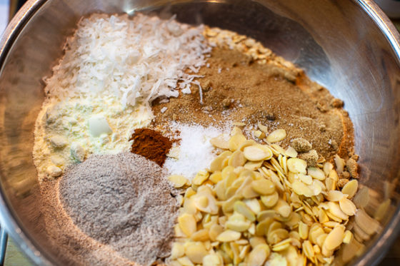 metal mixing bowl containing rolled oats, brown sugar, almonds, buckwheat, milk powder, salt, cinnamon, coconut flakes