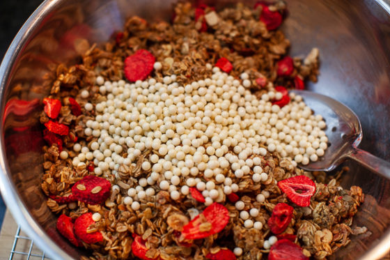 metal mixing bowl containing granola, dried strawberries and crunchy white chocolate pearls, being mixed with spoon