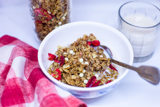 bowl of granola with spoon, next to red and white checkered napkin, jar of granola and small glass of milk.