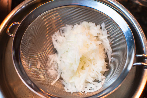 grated onion sitting in sieve over metal mixing bowl.