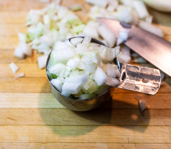 One cup measuring cup containing diced white onion on cutting board.