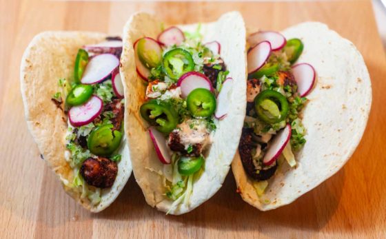 Three Octopus tacos with cabbage, jalapeno, radish, and fish sauce on a wood cutting board.